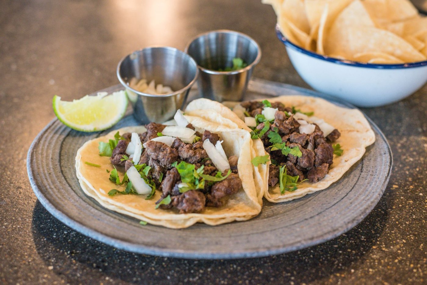 Two street tacos on corn tortillas with steak, onion and cilantro, served with lime and a bowl of tortilla chips at Mr. Tako.